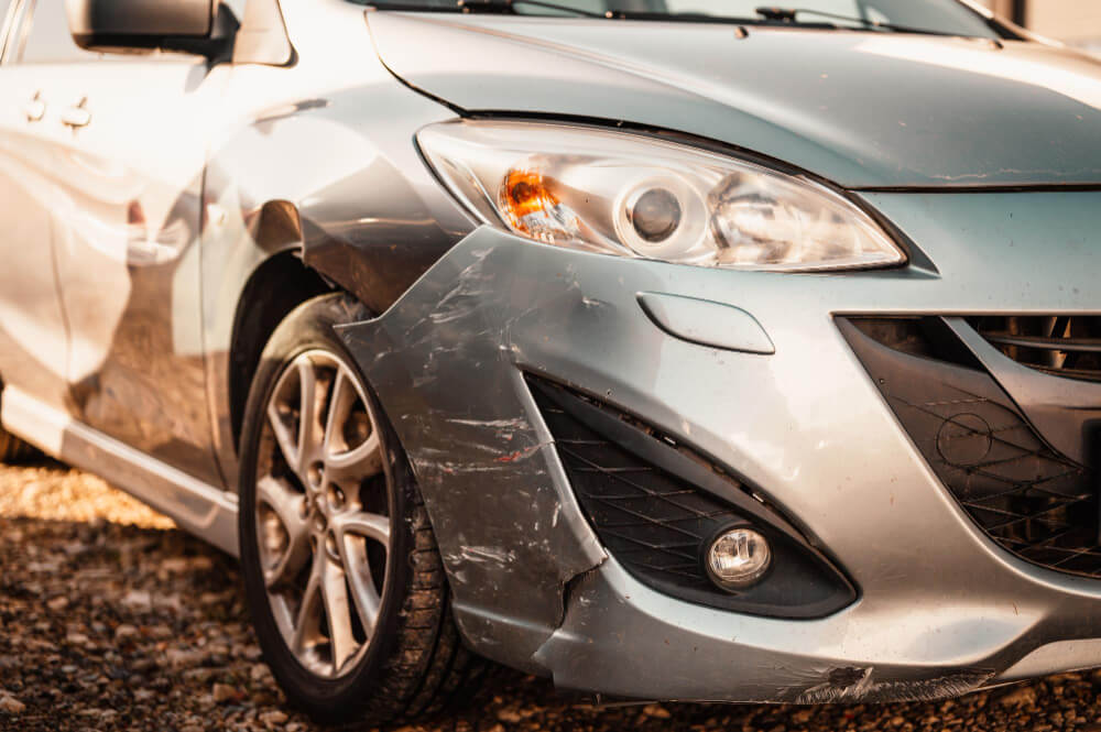 Nahaufnahme eines silbernen Autos mit sichtbaren Schäden und Kratzern am vorderen linken Stoßfänger und Kotflügel