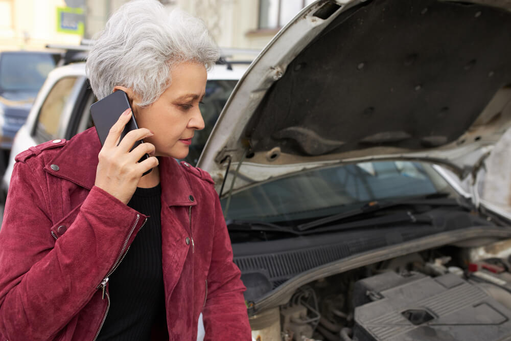 Eine Frau mit grauen Haaren im roten Jackett ruft um Hilfe neben einem Auto mit offener Motorhaube, möglicherweise für ein Unfallgutachten