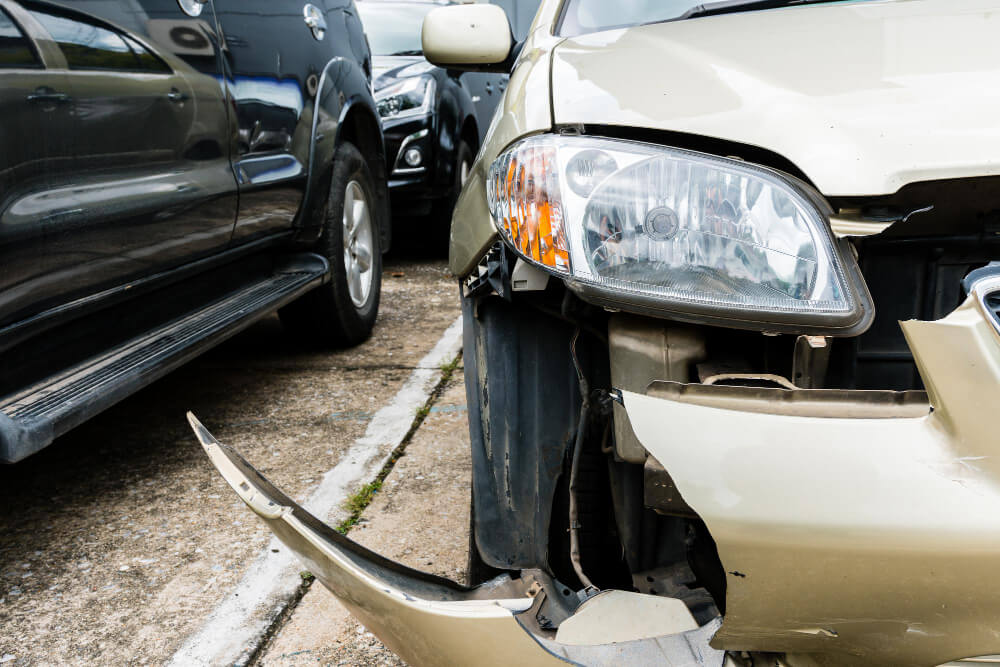 Ein beigefarbener Pkw mit beschädigter Frontstoßstange und Scheinwerfer steht neben einem schwarzen SUV auf einem Parkplatz für Schadengutachten