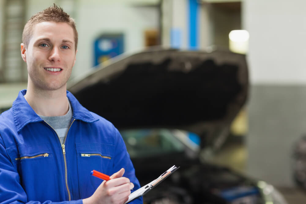 Ein lächelnder Mechaniker in blauer Arbeitskleidung mit Klemmbrett und Stift steht vor einem Auto mit offener Motorhaube in der Werkstatt