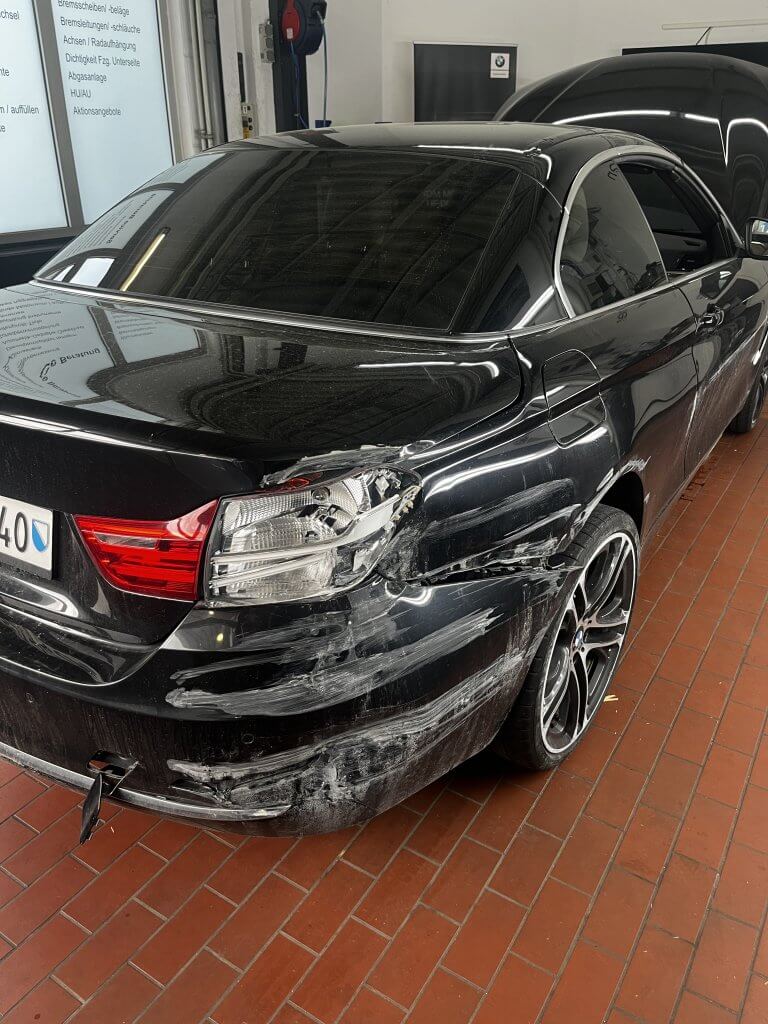 Ein schwarzes Auto mit deutlich sichtbaren Schäden und weißen Kratzern hinten links steht in einer Garage mit rotem Fliesenboden