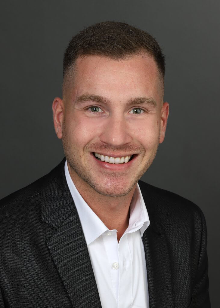 Ein lächelnder Mann im dunklen Anzug und weißen Hemd steht vor einem schlichten dunklen Hintergrund