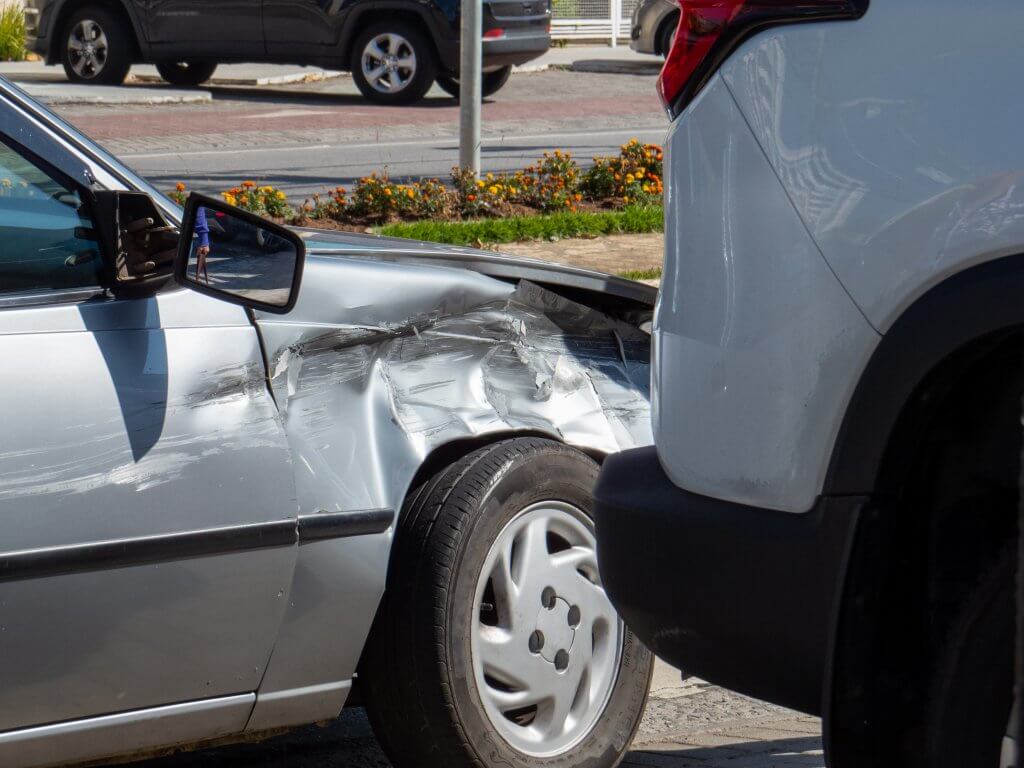Ein silbernes Auto mit eingedrückter Front und kaputtem Spiegel nach einem Unfall auf einer Stadtstraße, Automatisch gespeicherter Entwurf