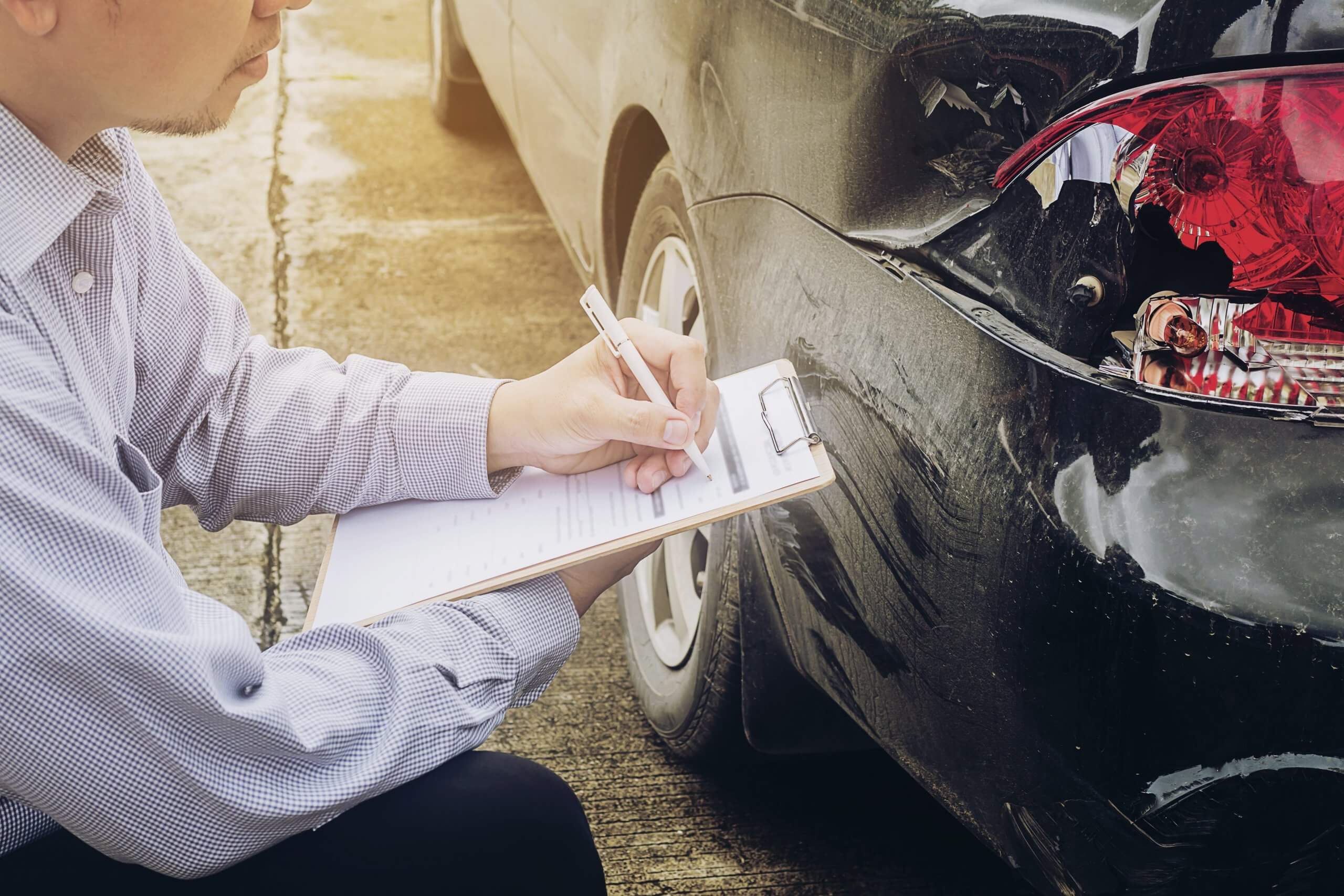 Eine Person schreibt auf ein Klemmbrett neben einem schwarzen Auto mit Heckschaden, vermutlich ein automatisch gespeicherter Unfallberichtsentwurf